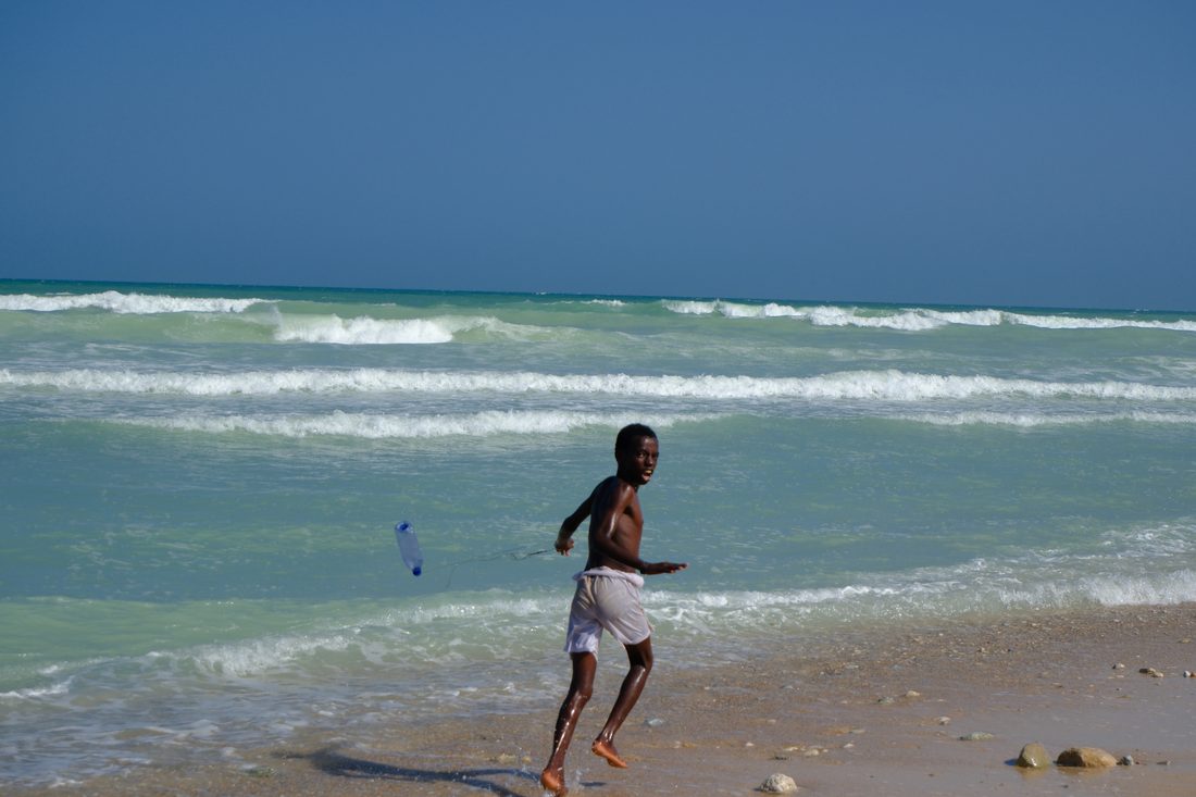 A boy runs along the shoreline throwing a plastic bottle on a line, with turquoise waves breaking behind him