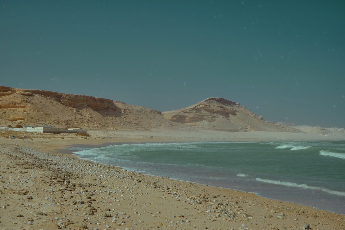 A wide empty beach of pale sand and pebbles curving beneath eroded sandstone cliffs, with turquoise surf breaking along the shore