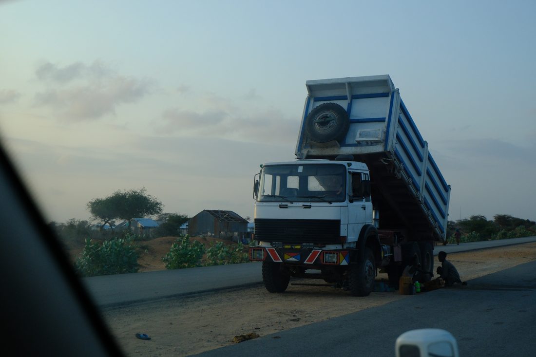 A white and blue tipper lorry stopped at the roadside with its bed raised while two people work underneath it